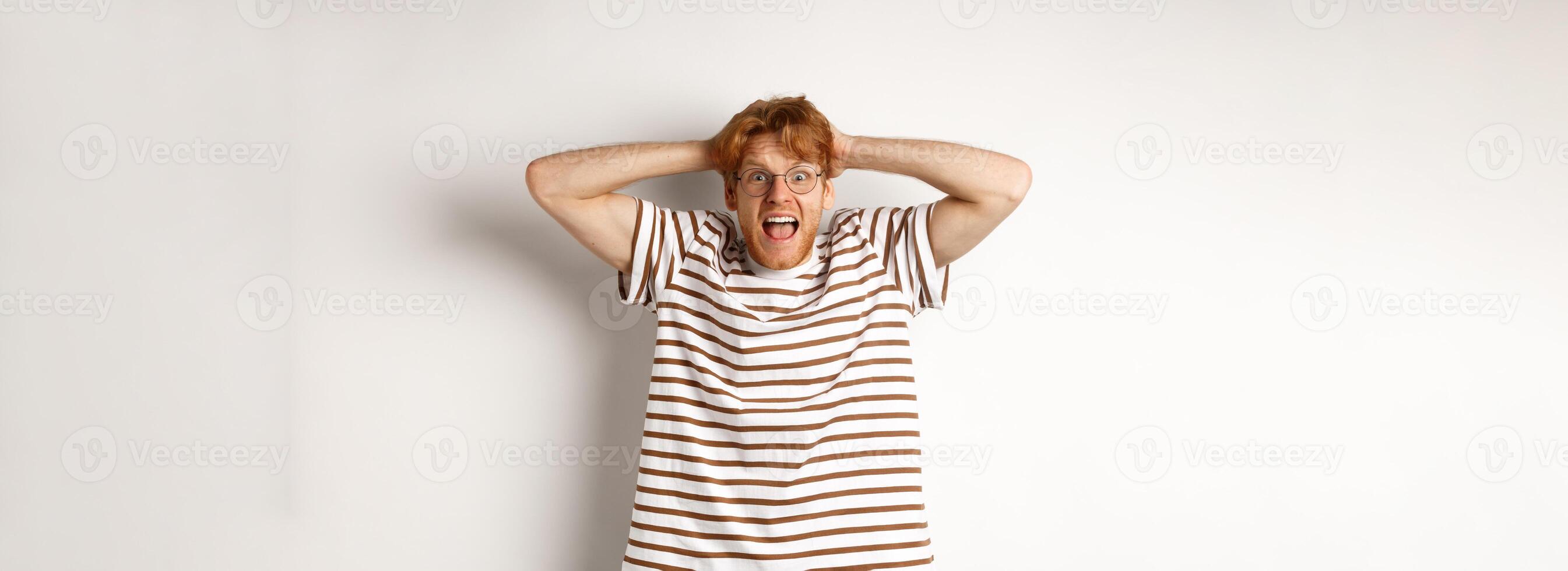 Frustrated redhead man panicking, screaming and holding hands on head, standing on white background photo
