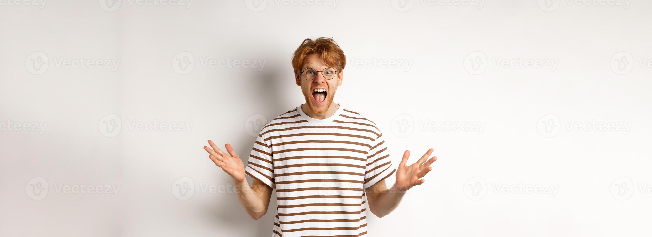 Frustrated man with red hair shaking hands and yelling at camera, looking angry and displeased, standing over white background photo