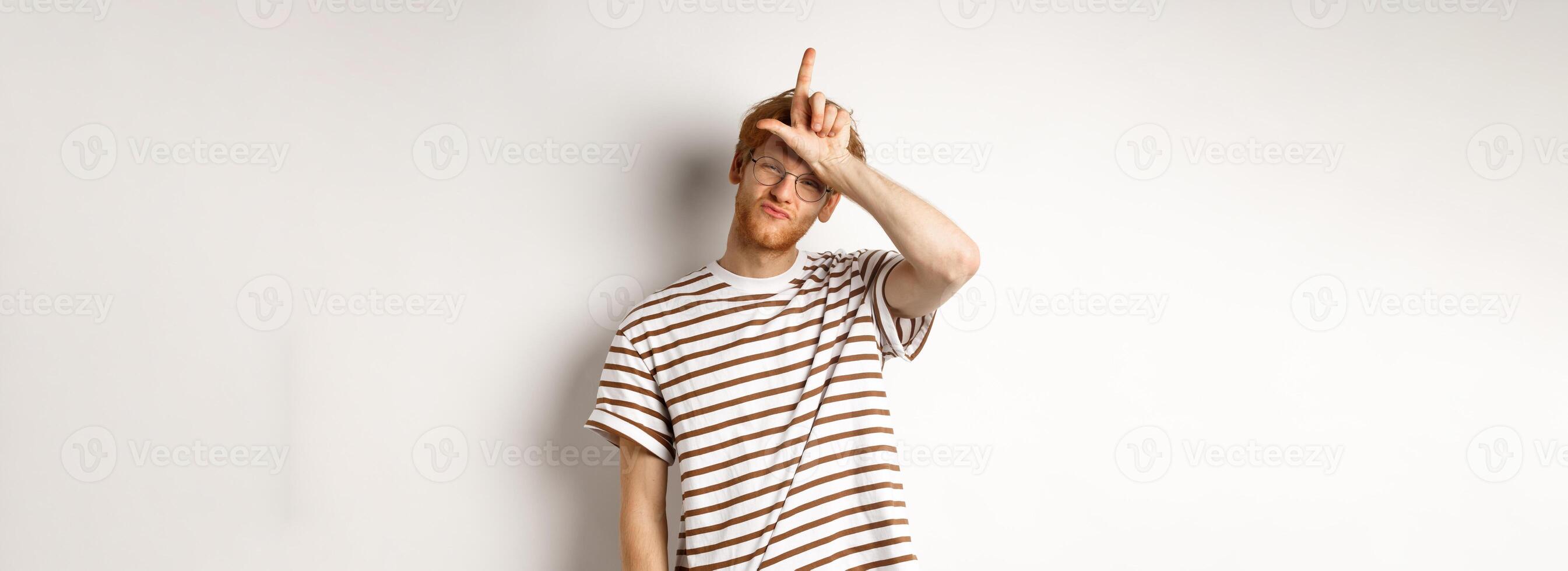 Sassy and arrogant redhead man scolding lost team, showing loser gesture on forehead and making smug face, standing over white background photo