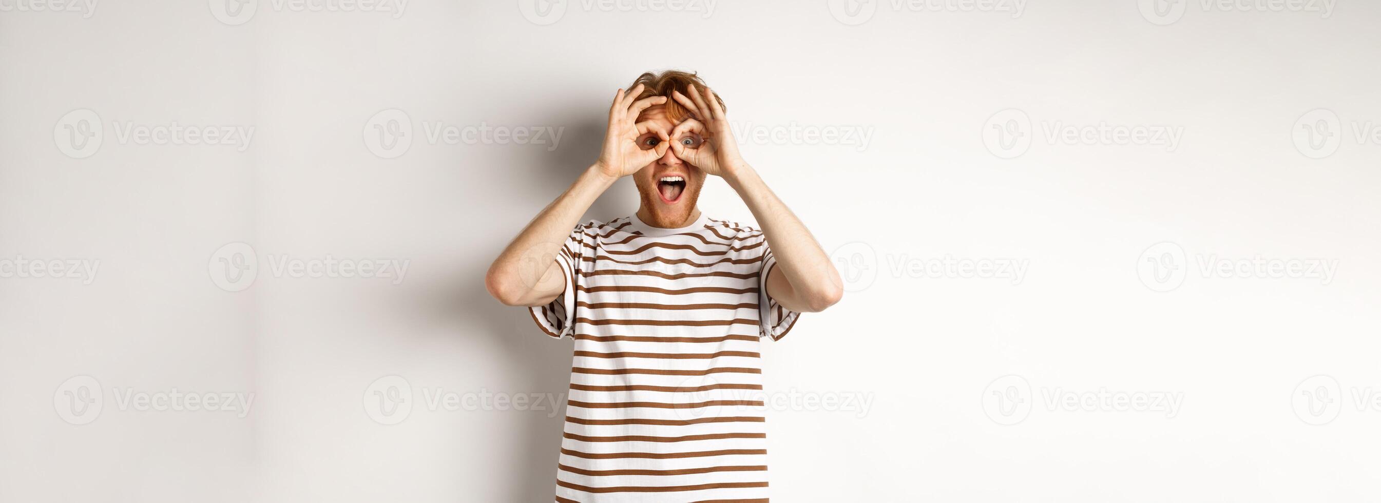 Impressed redhead man checking out promo offer, looking at camera from hand binoculars, smiling amazed, white background photo
