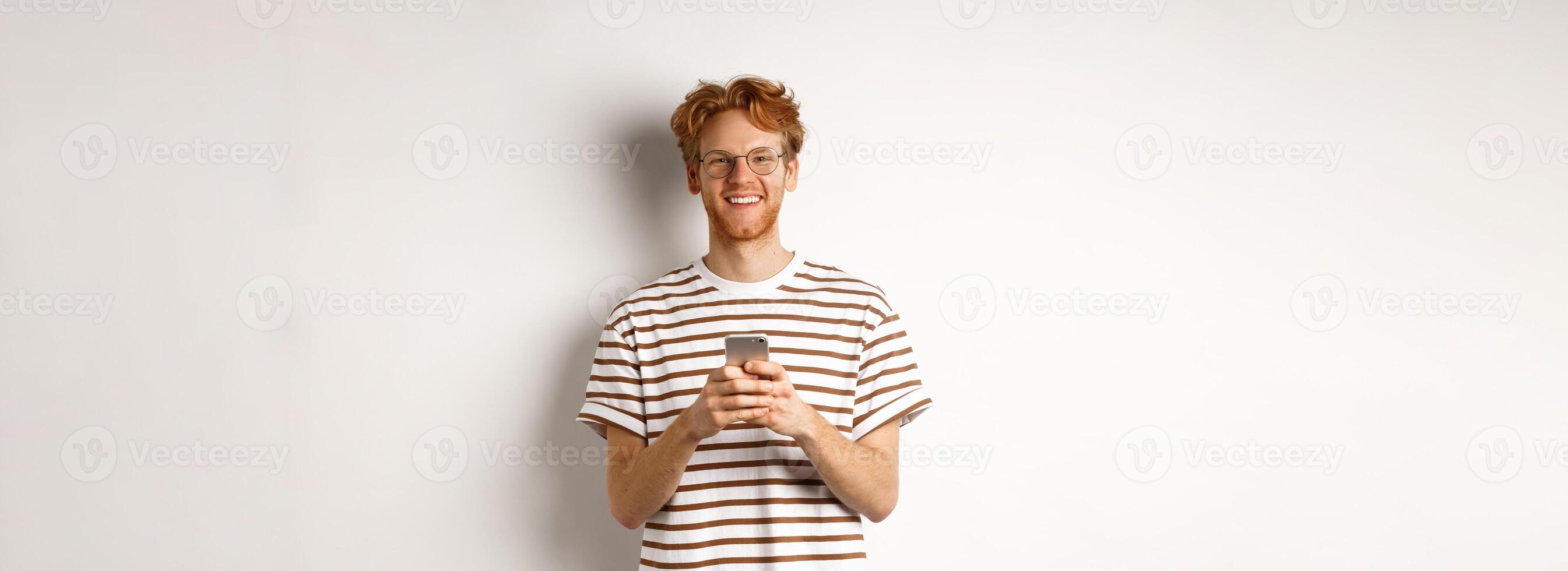 Technology and e-commerce concept. Redhead guy in glasses using mobile phone and smiling. Young man with smartphone staring happy at camera, white background photo