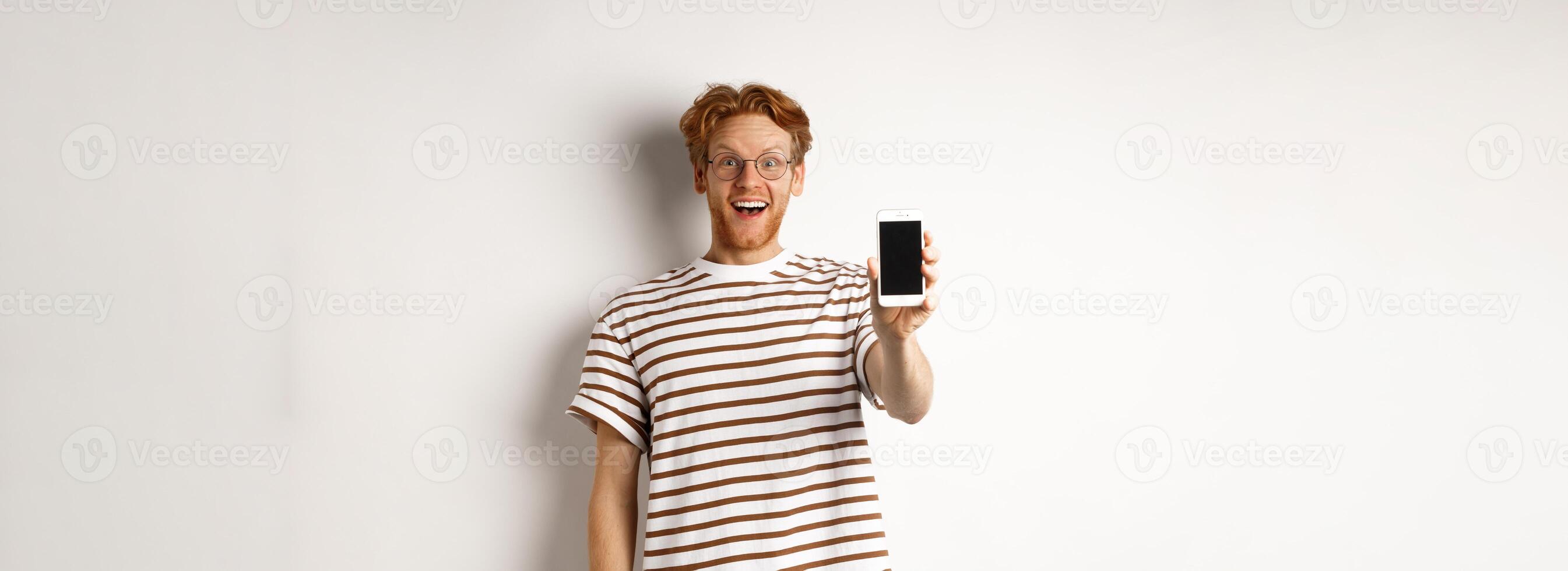Technology and e-commerce concept. Happy young redhead man in glasses showing blank smartphone screen, looking at camera amazed, standing over white background photo