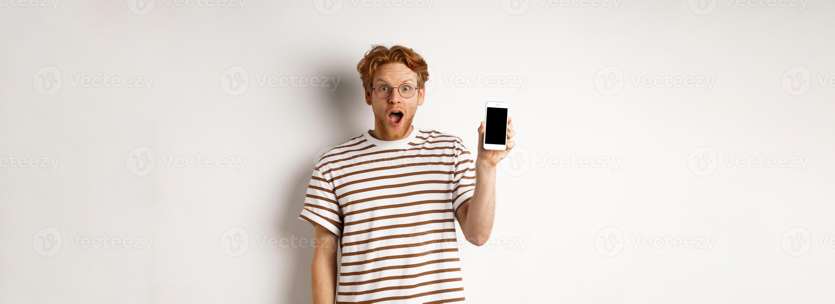 Technology and e-commerce concept. Surprised and shocked redhead guy checking out online promotion, showing blank smartphone screen and drop jaw, white background photo