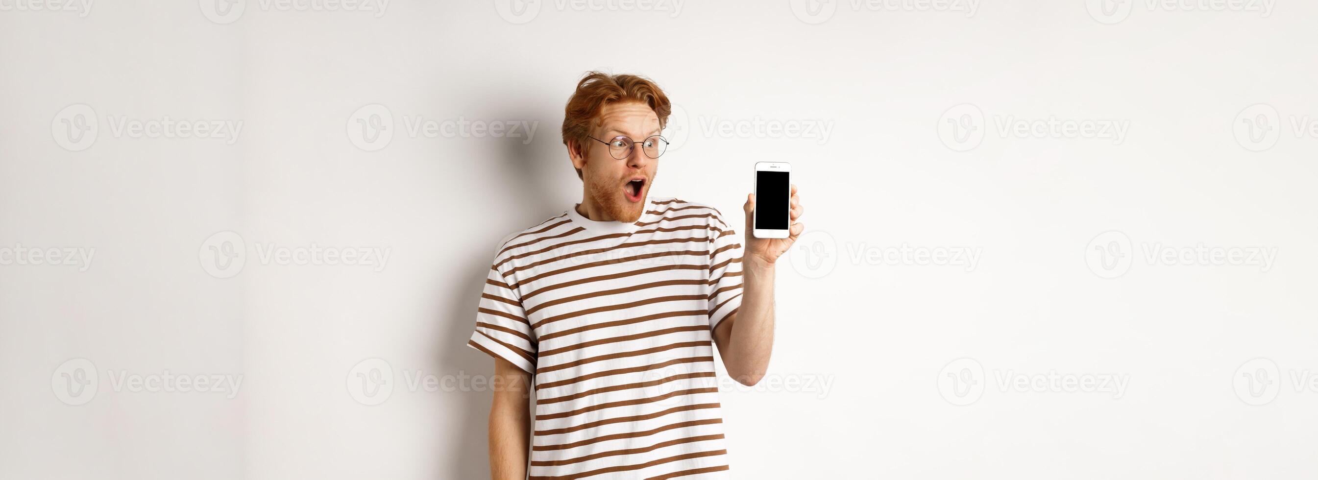 Technology and e-commerce concept. Surprised and shocked redhead guy checking out online promotion, showing blank smartphone screen and drop jaw, white background photo