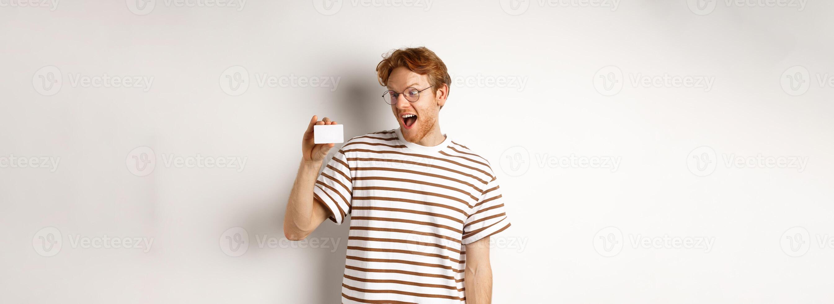 Shopping and finance concept. Excited young man with red hair, staring at plastic card and scream of joy, standing over white background photo