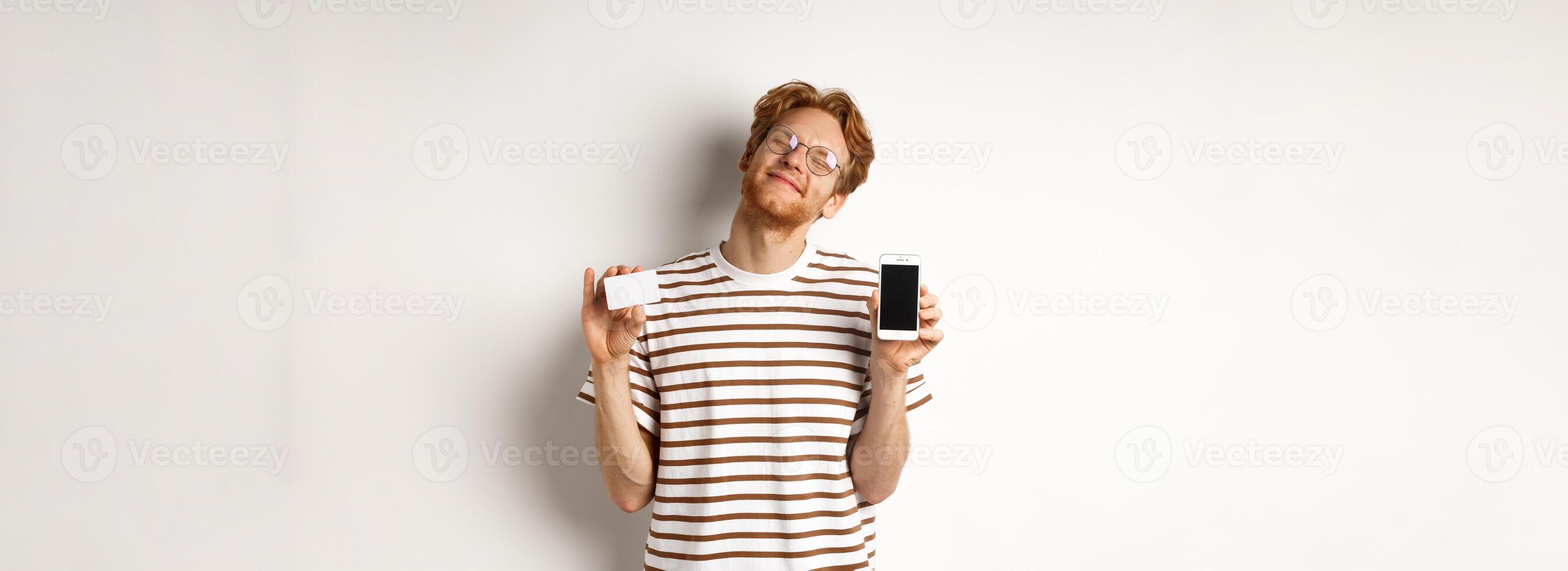 Shopping and finance concept. Pleased young man with red hair smiling from satisfaction, showing smartphone blank screen and credit card, white background photo