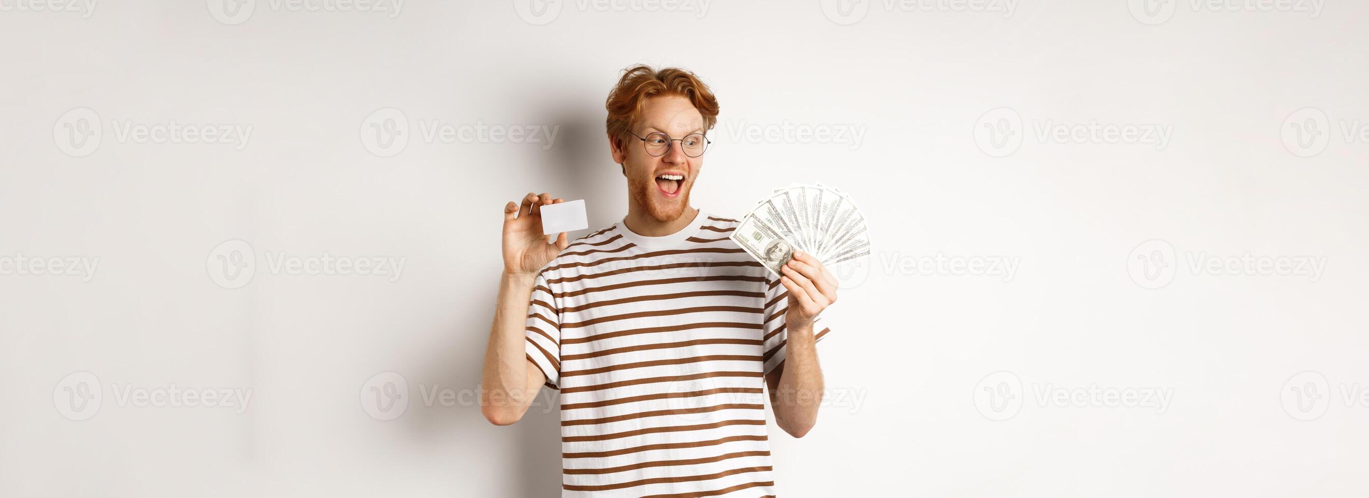 Shopping and finance concept. Cheerful handsome redhead man showing plastic credit card and cash, smiling amazed, white background photo