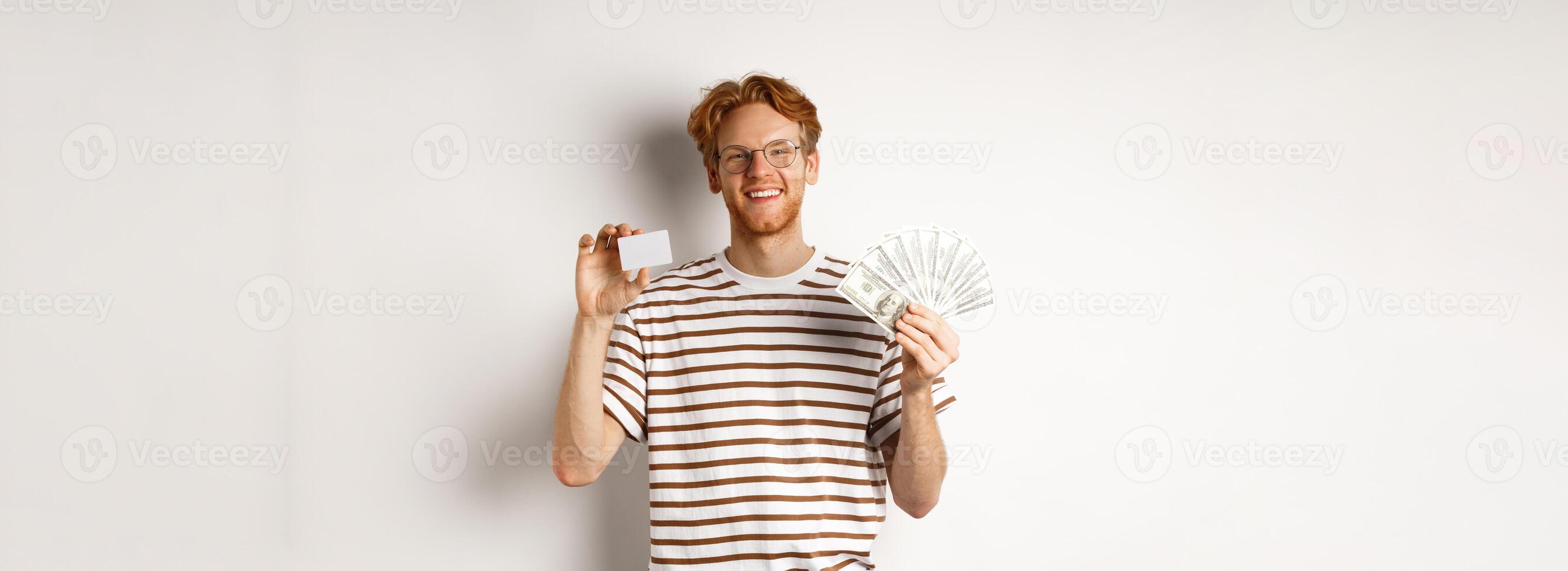Shopping and finance concept. Young redhead man with beard and glasses showing plastic credit card with money in dollars, white background photo