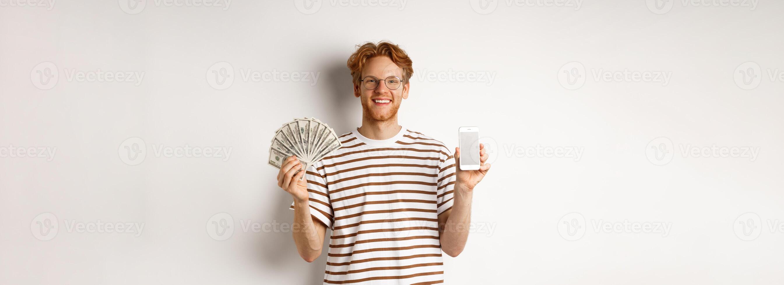Smiling young redhead man in glasses showing smartphone blank screen and money, standing over white background photo