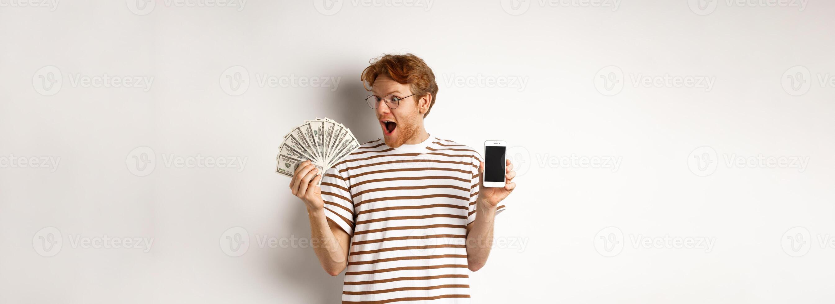 Amazed redhead man showing smartphone app on blank screen and money, winning prize cash online, standing over white background photo