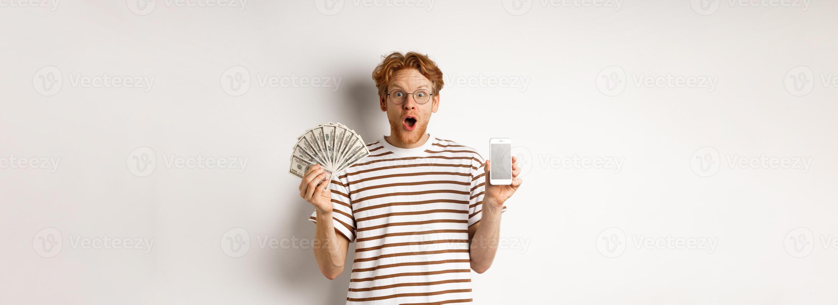 Amazed redhead man showing smartphone app on blank screen and money, winning prize cash online, standing over white background photo