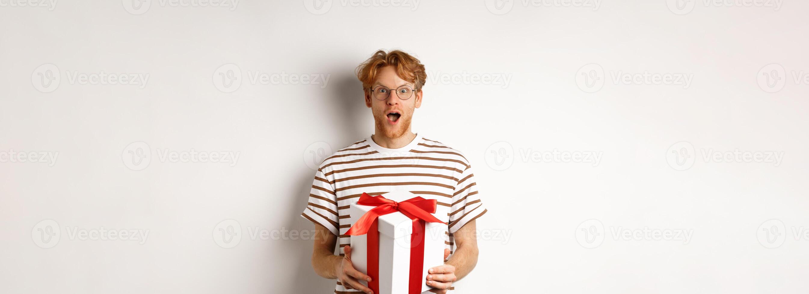 Valentines day and holidays concept. Surprised redhead boyfriend in glasses looking grateful at camera, receive big present in box, standing over white background photo