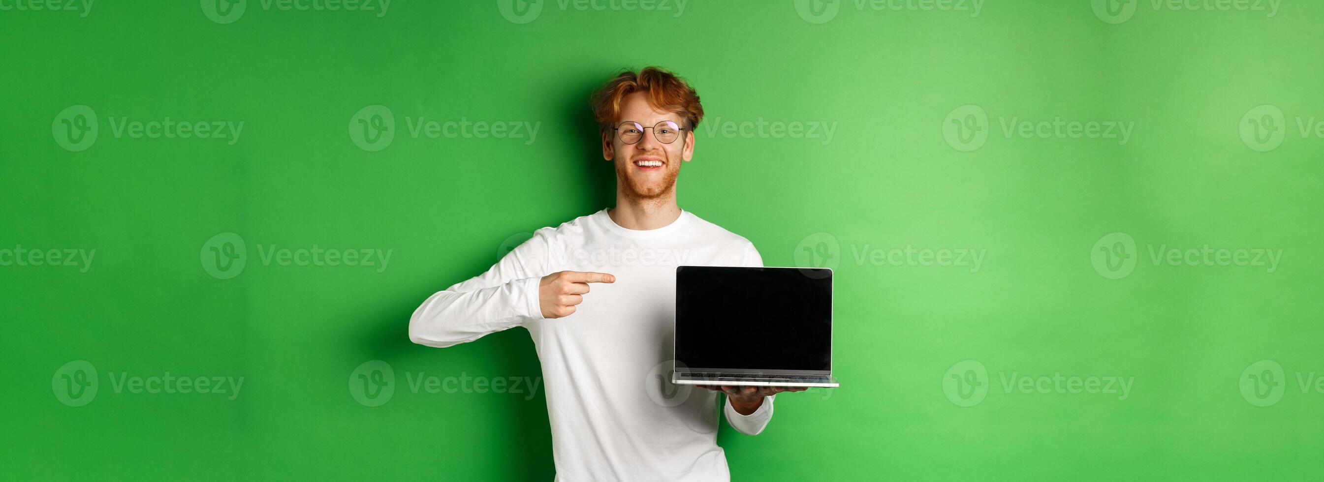 Happy redhead guy in glasses and white long-sleeve t-shirt, pointing finger at blank laptop screen and smiling, standing over green background photo