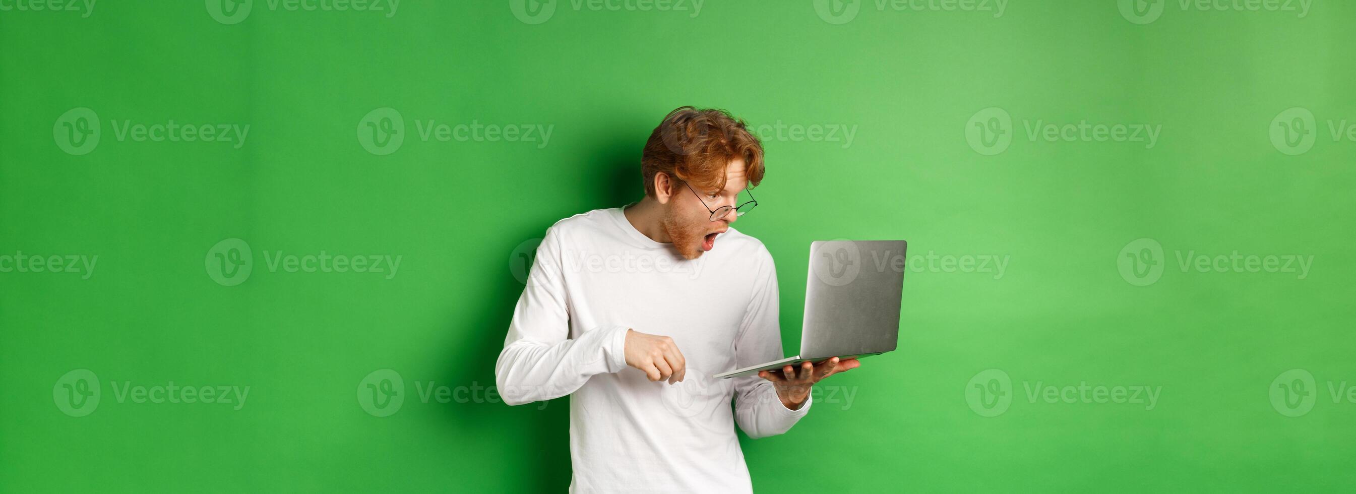 Image of young redhead man looking surprised at laptop screen, drop jaw and staring at display amazed, green background photo