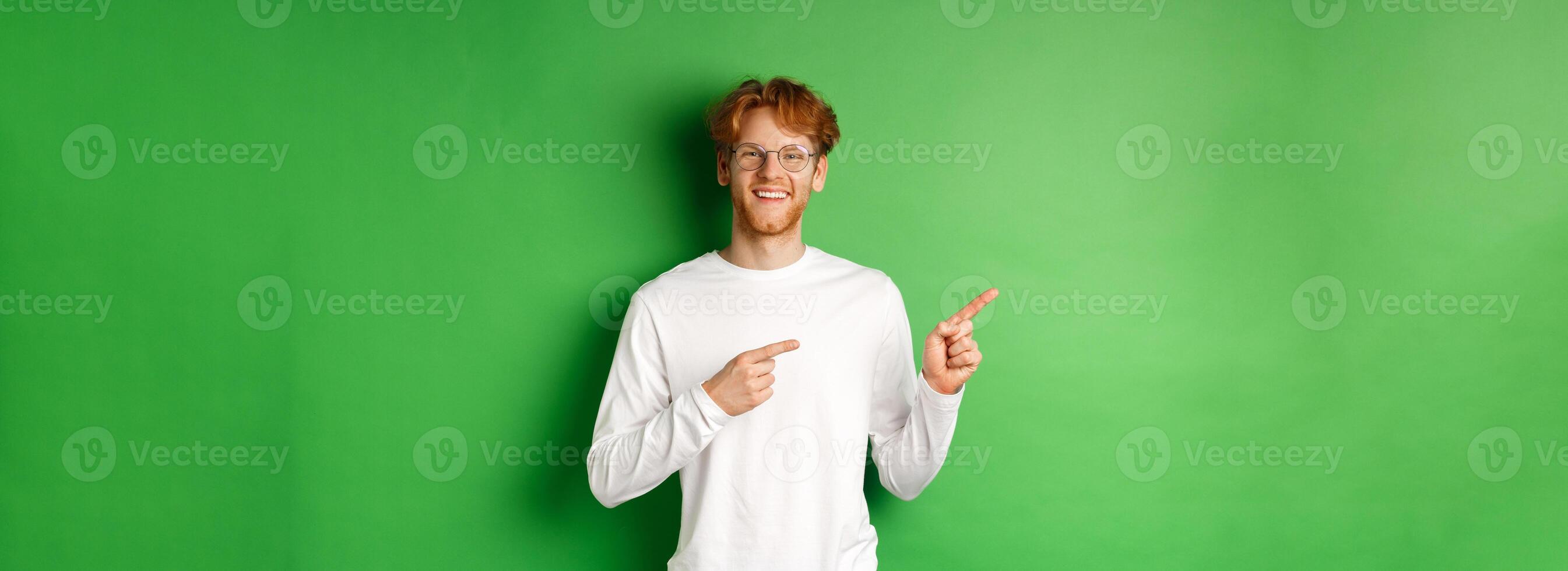 Handsome young man with red hair and beard smiling, pointing fingers left at logo, showing advertisement, standing over green background photo