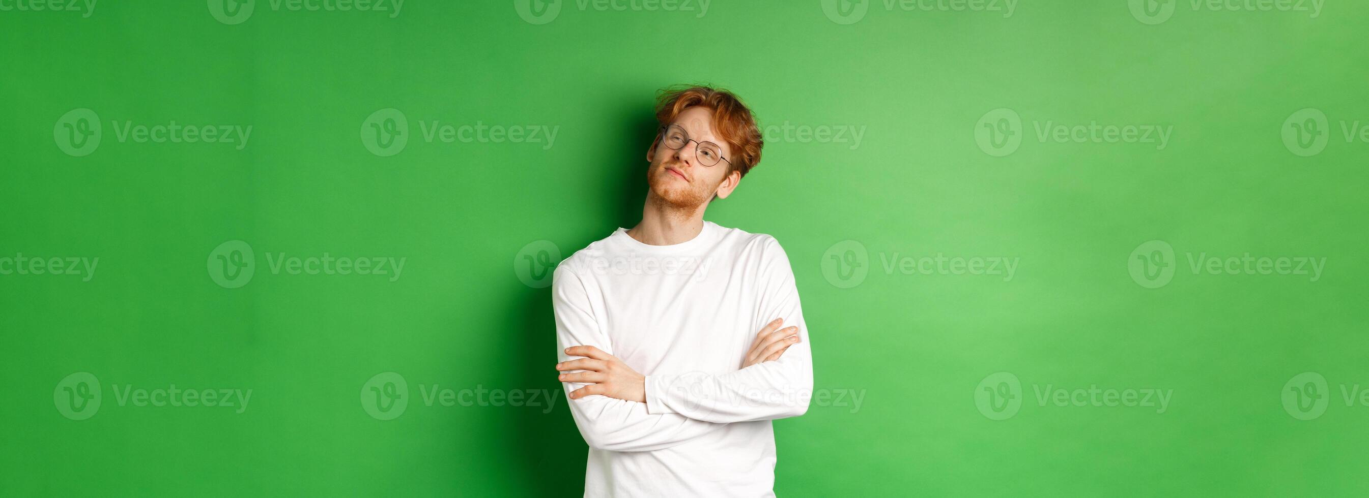 Confident young man with red hair, wearing glasses, looking smug at right side copy space, cross arms on chest self-assured, standing over green background photo