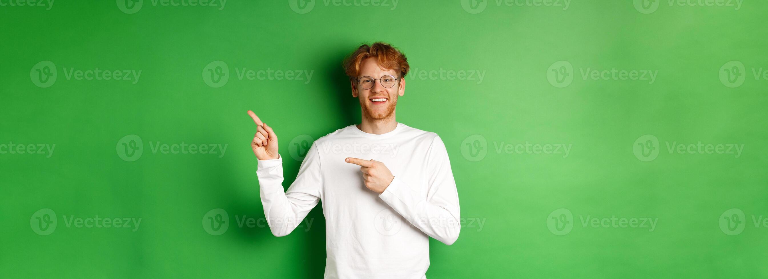 Handsome young man with ginger hair and glasses, pointing fingers right at copy space and smiling, standing over green background photo
