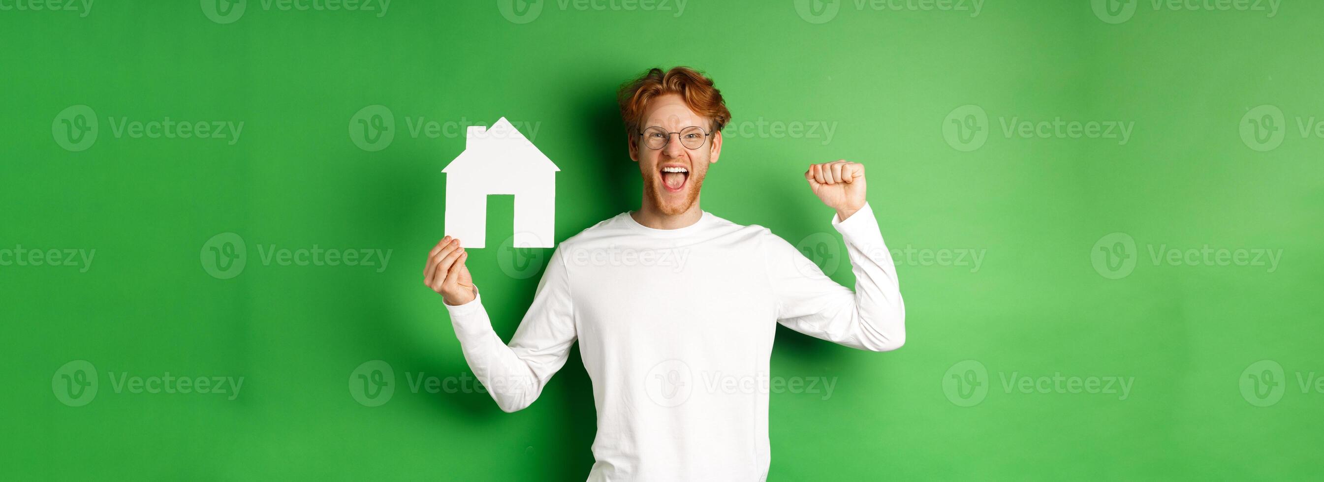 Real estate. Happy caucasian man in glasses with red hair celebrating, showing paper house cutout and making fist pump sign, shouting for joy, buying property, green background photo