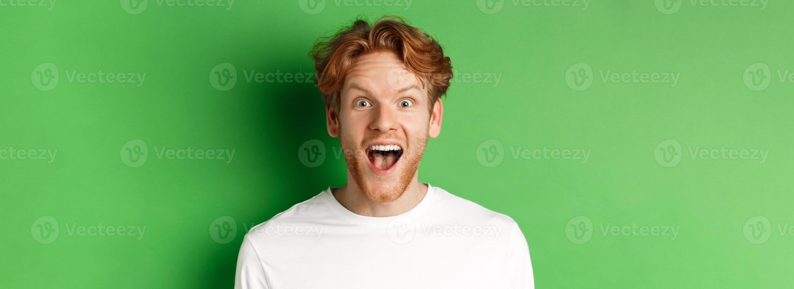 Emotions and fashion concept. Close up of surprised redhead man hear amazing news, staring at camera joyful, standing over green background photo