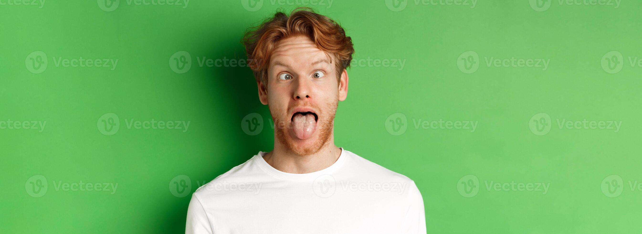 Emotions and fashion concept. Close up of funny redhead man showing silly faces, sticking tongue and squinting, standing over green background photo