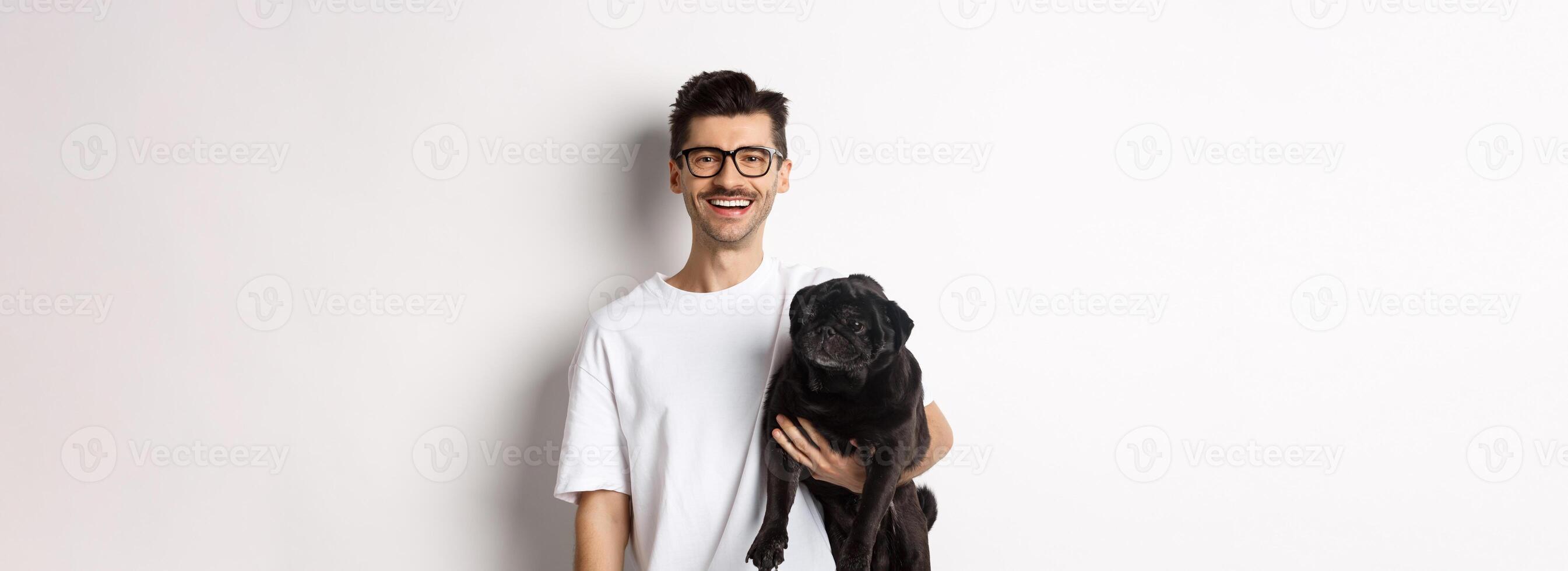 Handsome hipster guy holding his funny black pug dog, smiling at camera, standing over white background photo