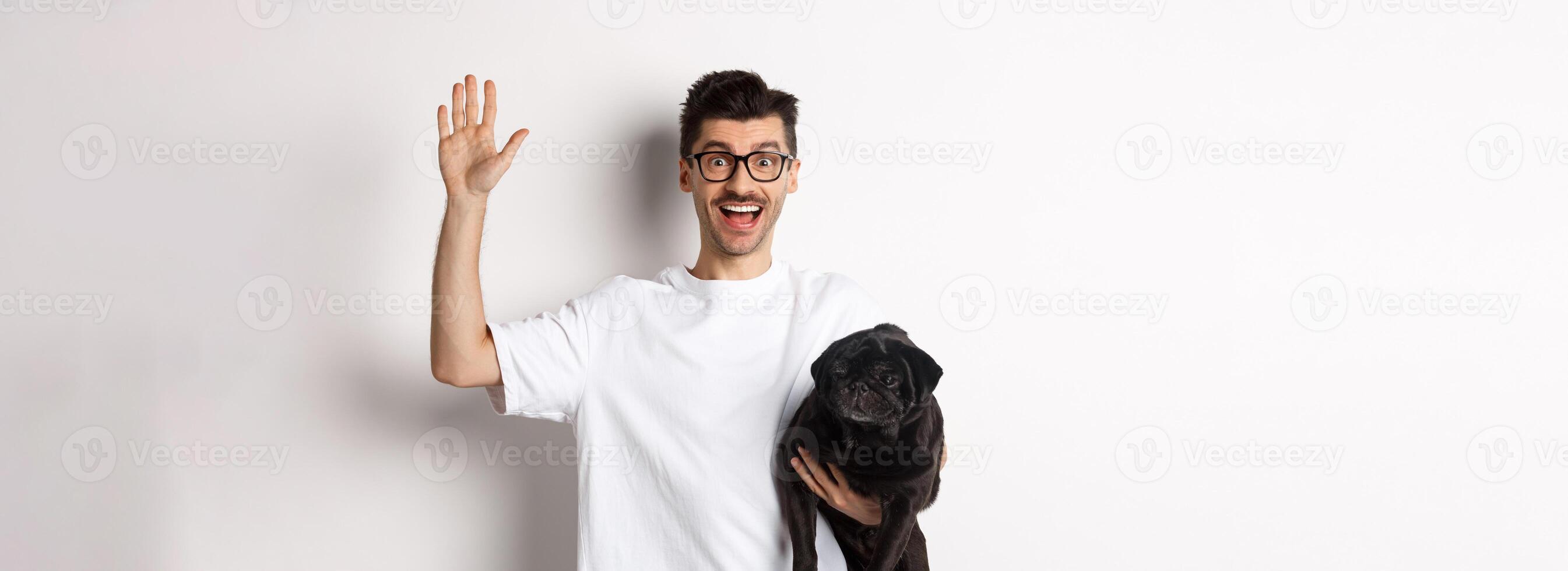 Handsome young man in glasses holding his black pug and waving hand, guy saying hello while carry dog with one arm, standing over white background photo