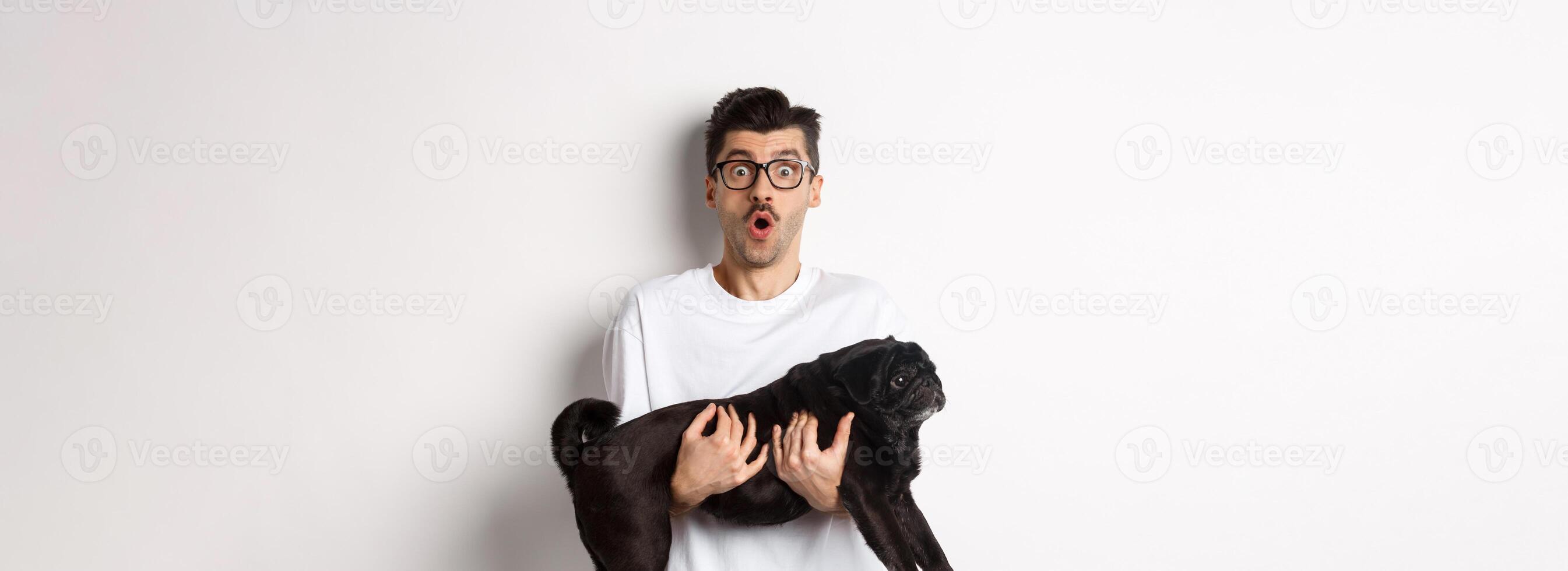 Surprised young man in glasses holding cute black pug, dog owner staring at camera with impressed face, saying wow, standing over white background photo