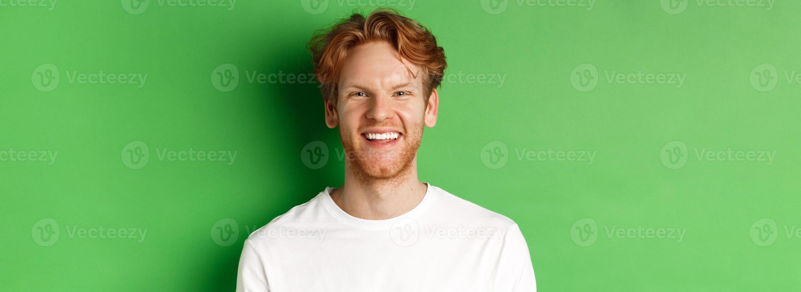 Emotions and fashion concept. Happy young man with red hair and beard, smiling and laughing at camera, standing over green background photo