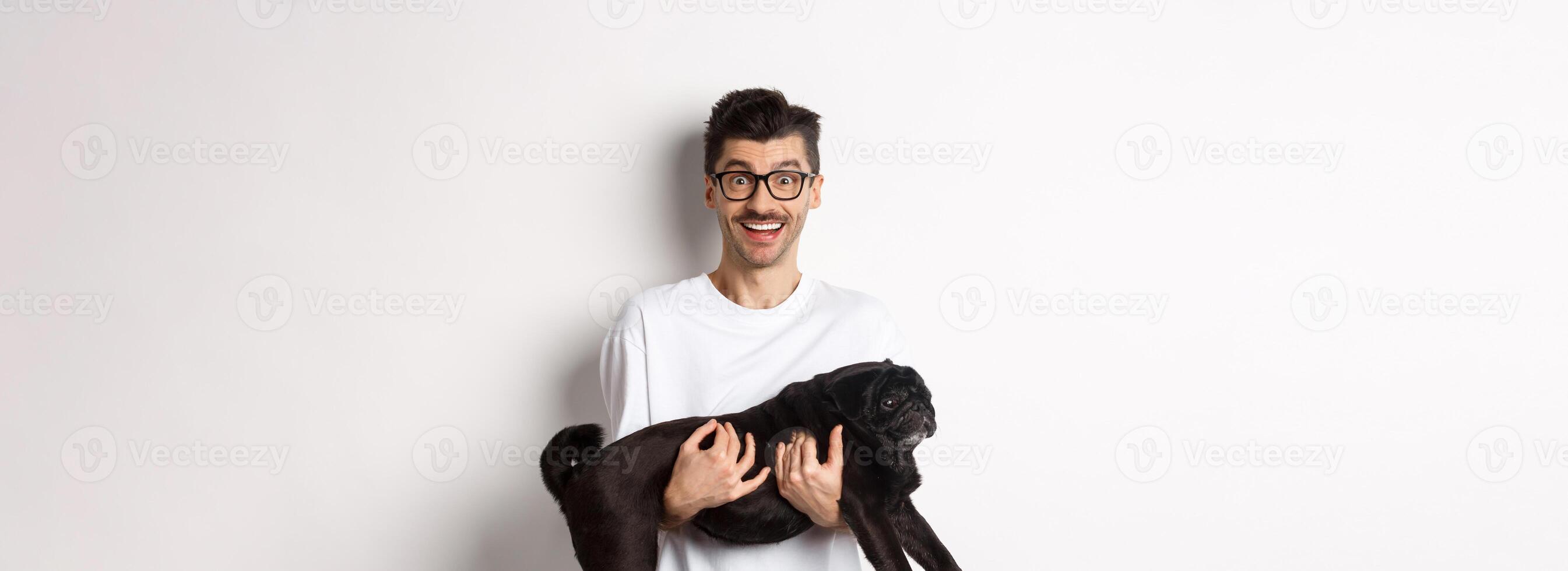 Happy young man in glasses holding black pug pet and staring at camera amused, smiling cheerful, standing over white background photo