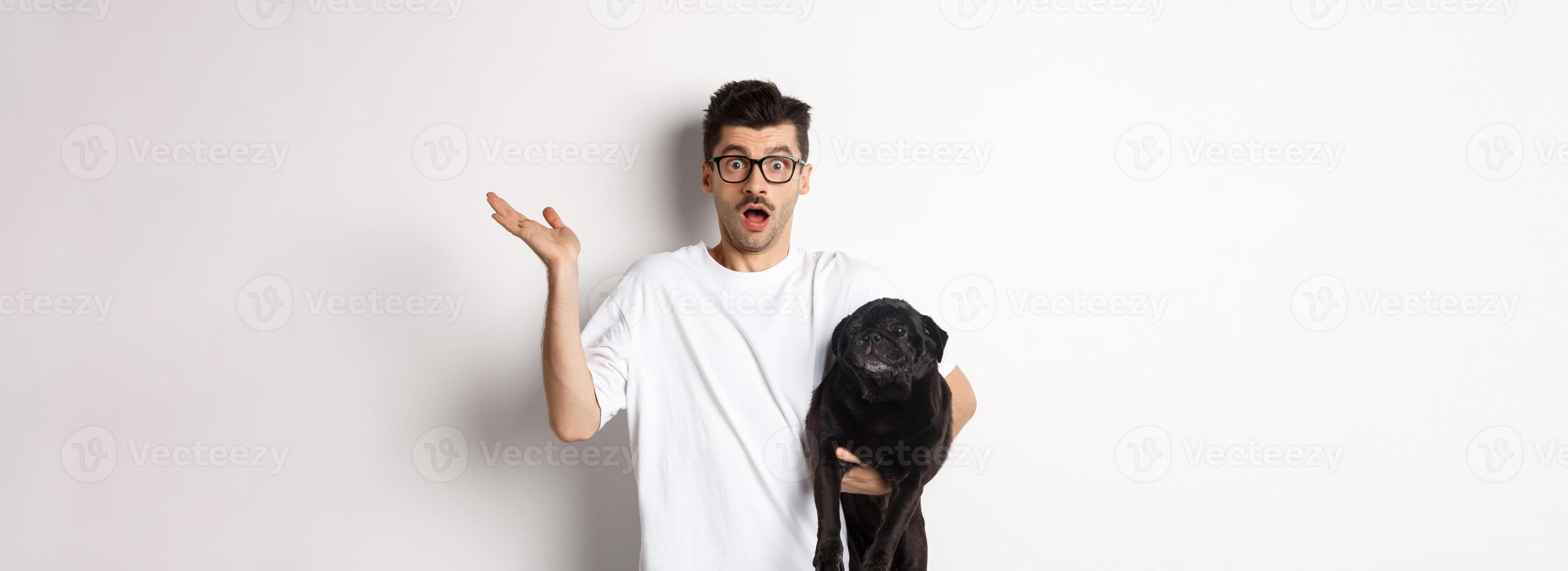 Image of confused hipster man holding dog and shrugging, dont know, raising hand puzzled, standing with his animal over white background photo
