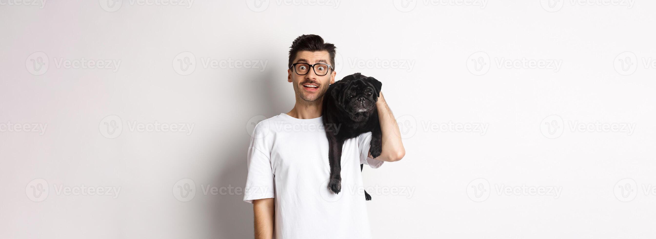 Amazed young man in glasses holding black pug on shoulder and staring at camera impressed. Dog owner posing with cute puppy near white background photo