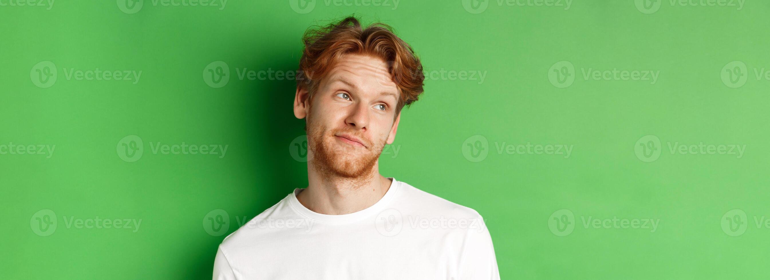Smiling redhead man with messy haircut and beard tilt head, looking left with pleased face, standing over green background photo