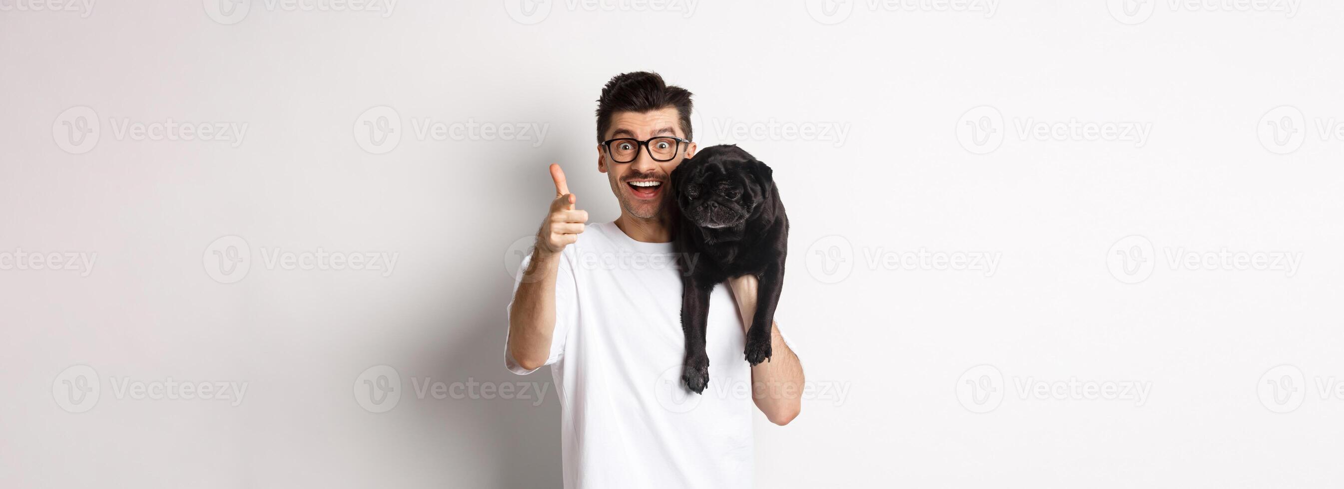 Happy young man holding cute black dog on shoulder and pointing at camera. Hipster guy carry pug on shoulder and staring at camera excited, standing over white background photo