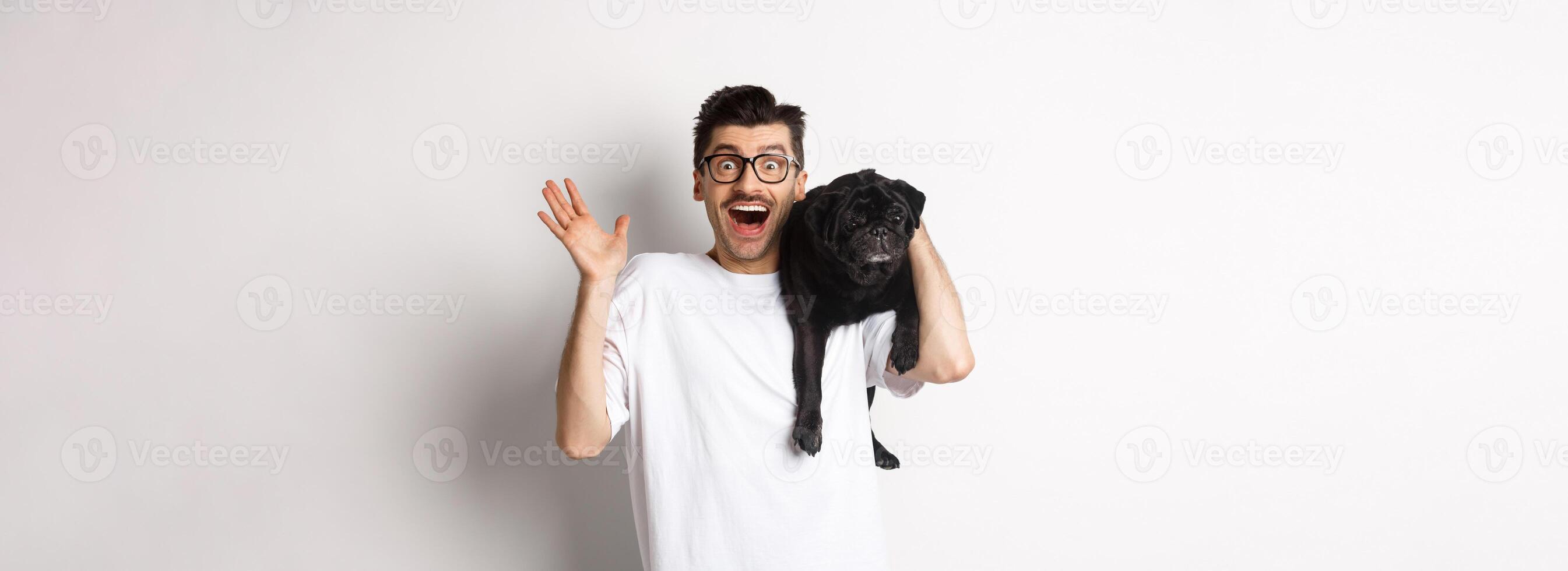 Excited hipster guy in glasses holding dog on shoulder, waving hand to say hello, greeting someone with happy face. Happy man carry his cute pug and smiling, white background photo