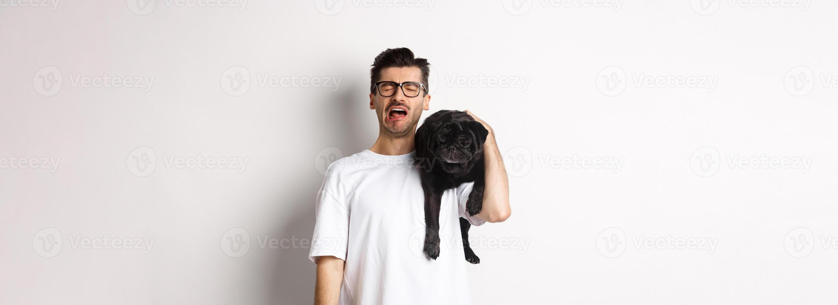 Sad dog owner crying, holding cute black pug on shoulder and looking miserable, sobbing while standing over white background photo
