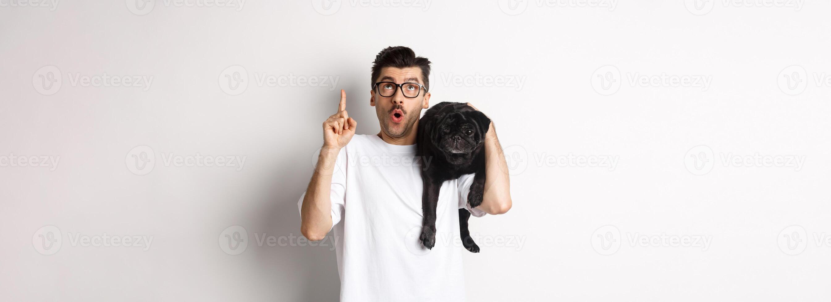 Amazed handsome man in glasses, holding cute black pug dog on shoulder, pointing finger up at promo logo, standing over white background photo