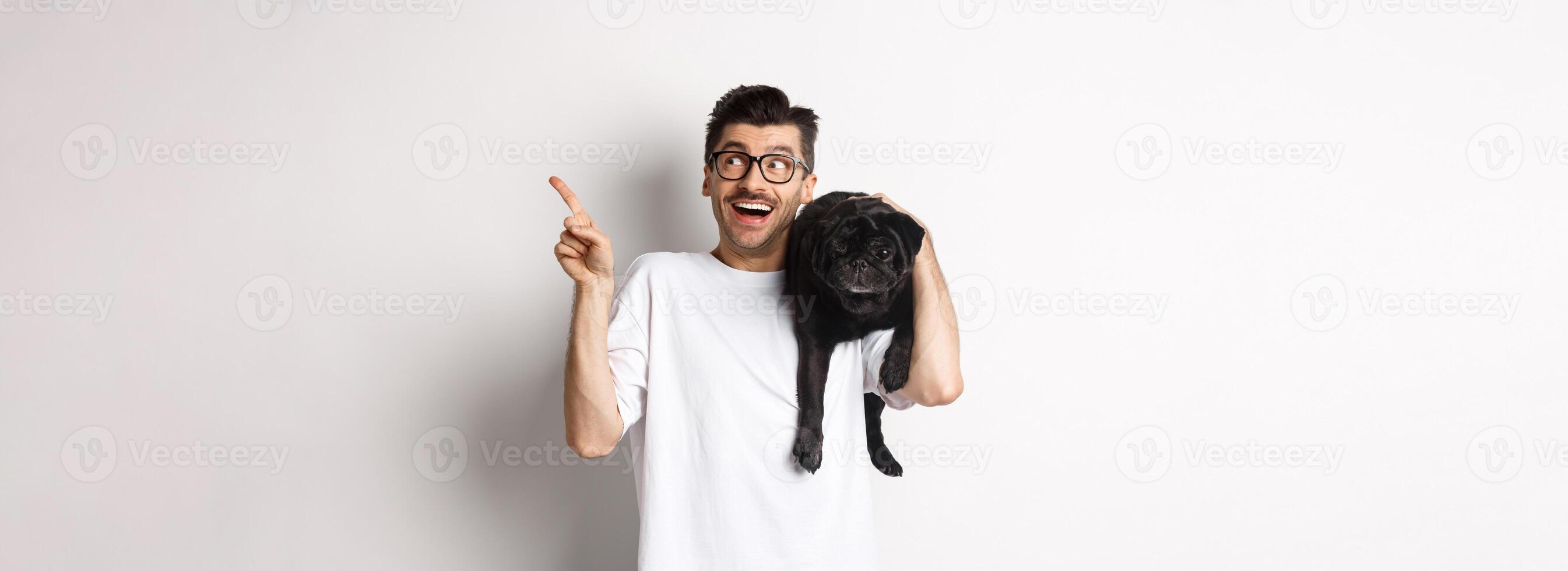 Amazed handsome man in glasses, holding cute black pug dog on shoulder, pointing finger upper right corner at promo logo, standing over white background photo