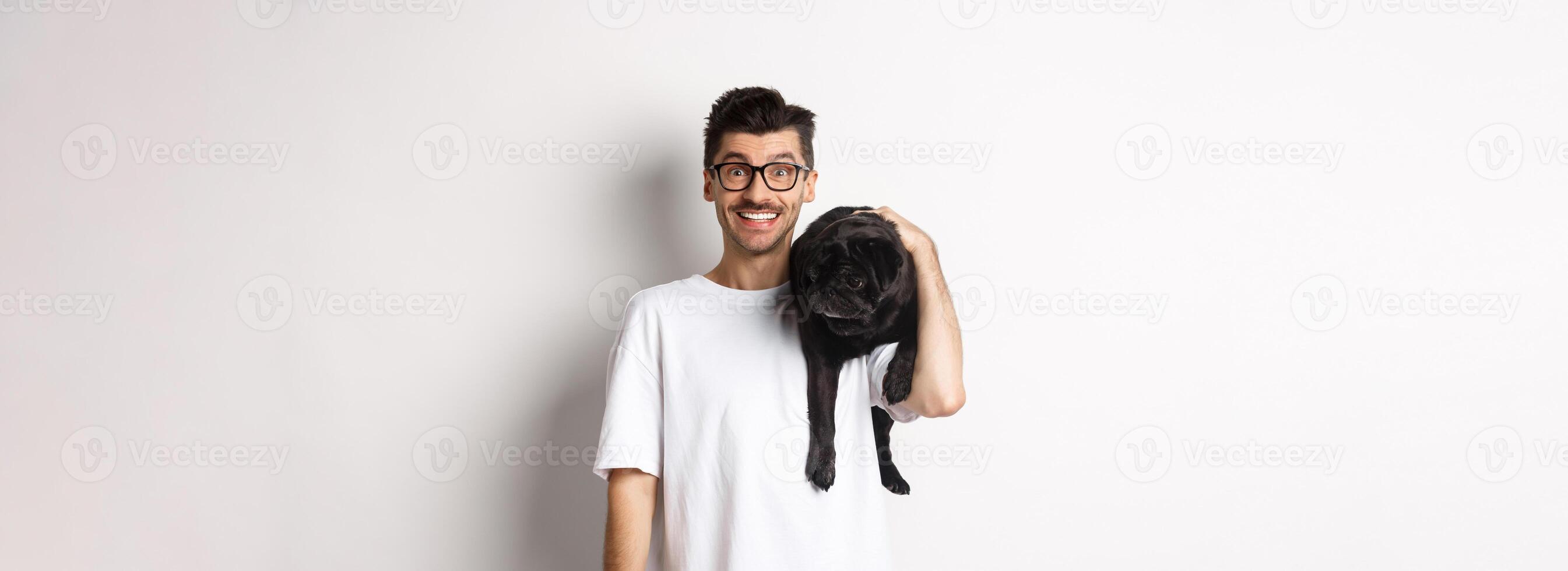 Cheerful hipster man in glasses, holding cute black pug dog on shoulder, staring at camera with happy smile, standing over white background photo