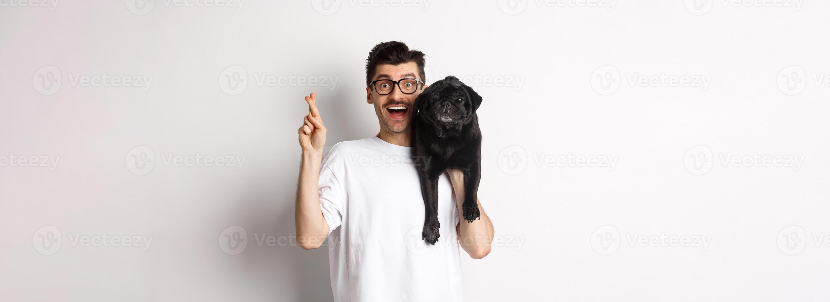 Hopeful smiling dog owner making a wish, holding cute black pug on shoulder and cross fingers for good luck, white background photo