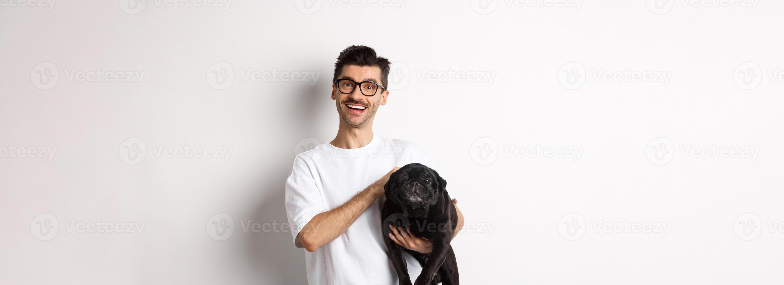 Happy hipster guy in glasses pet dog and smiling. Cute black pug enjoy spending time with owner, looking satisfied, standing over white background photo