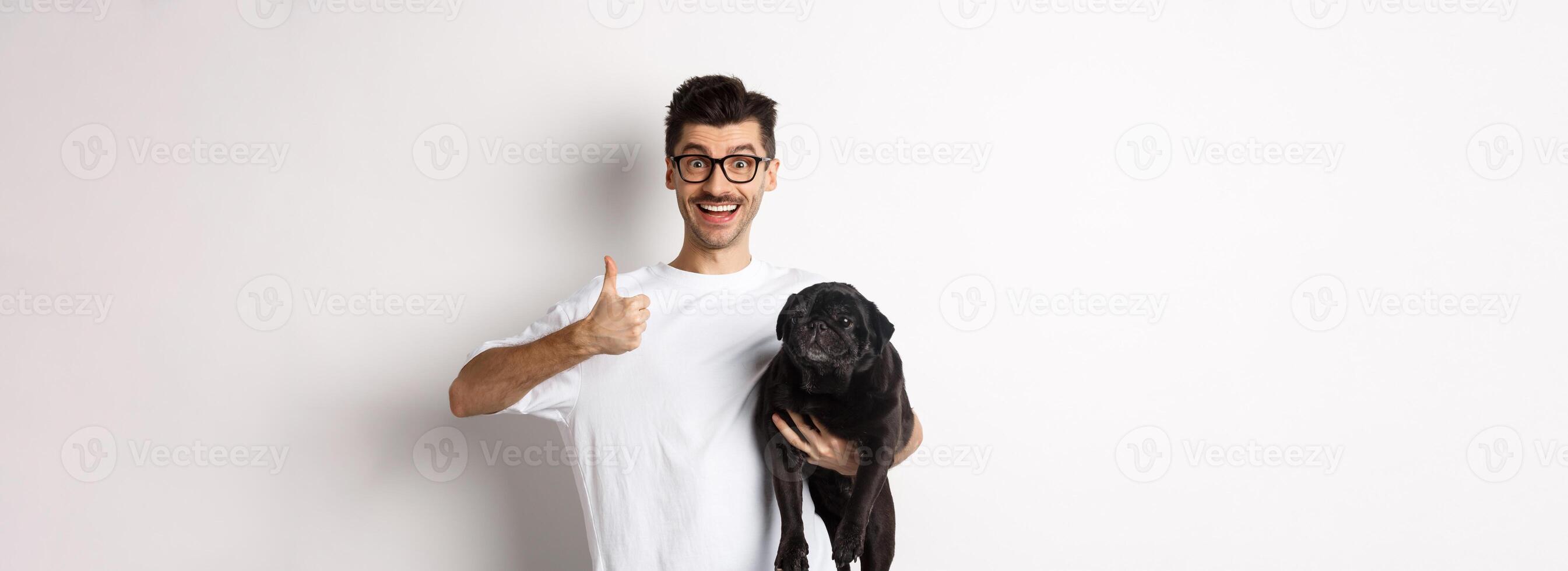 Cheerful handsome man holding dog and showing okay sign, approve or recommend product. Hipster guy carry cute black pug and looking satisfied, white background photo