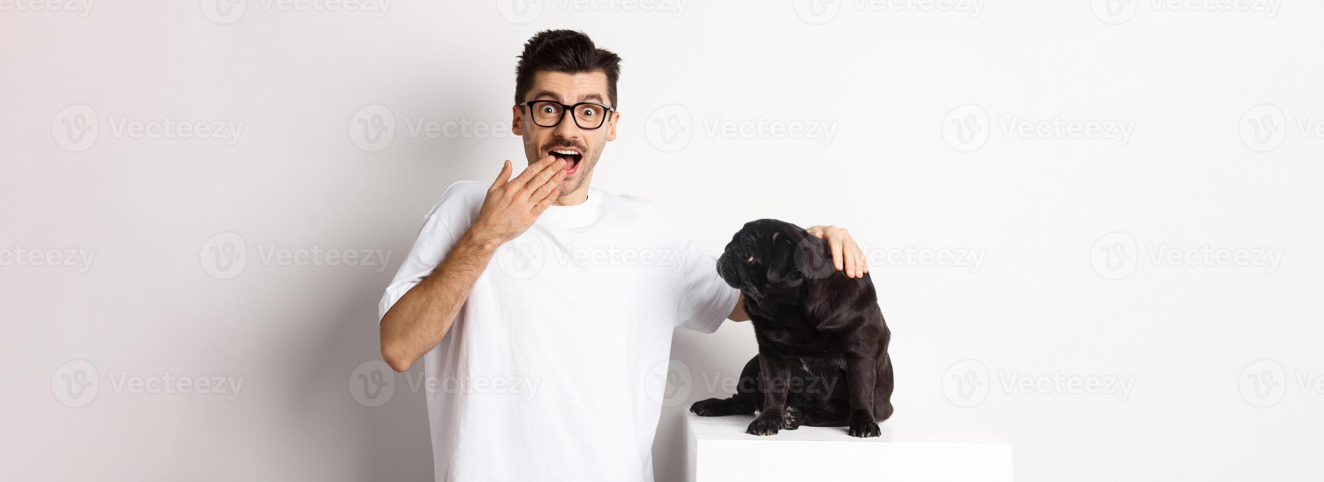 Image of surprised and happy dog owner pet his dog and staring at camera with rejoice, standing with pug over white background photo