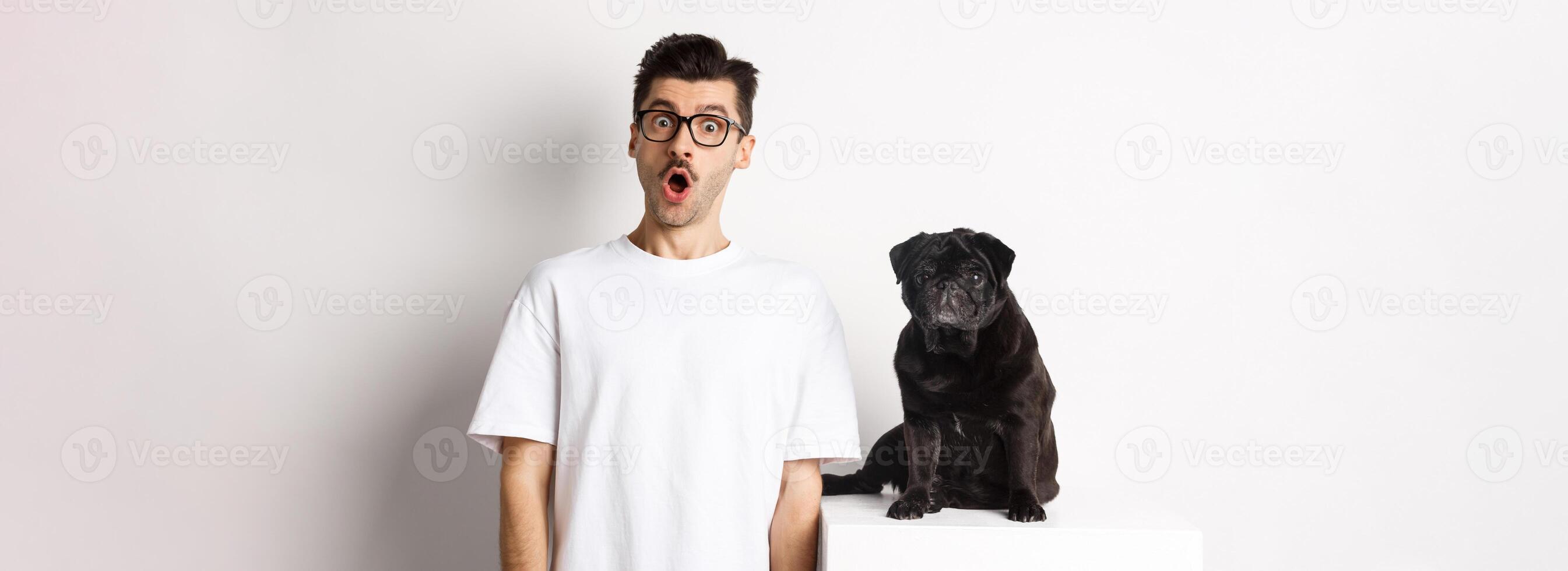Image of dog owner and cute black pug looking at camera surprised and amazed, standing over white background photo