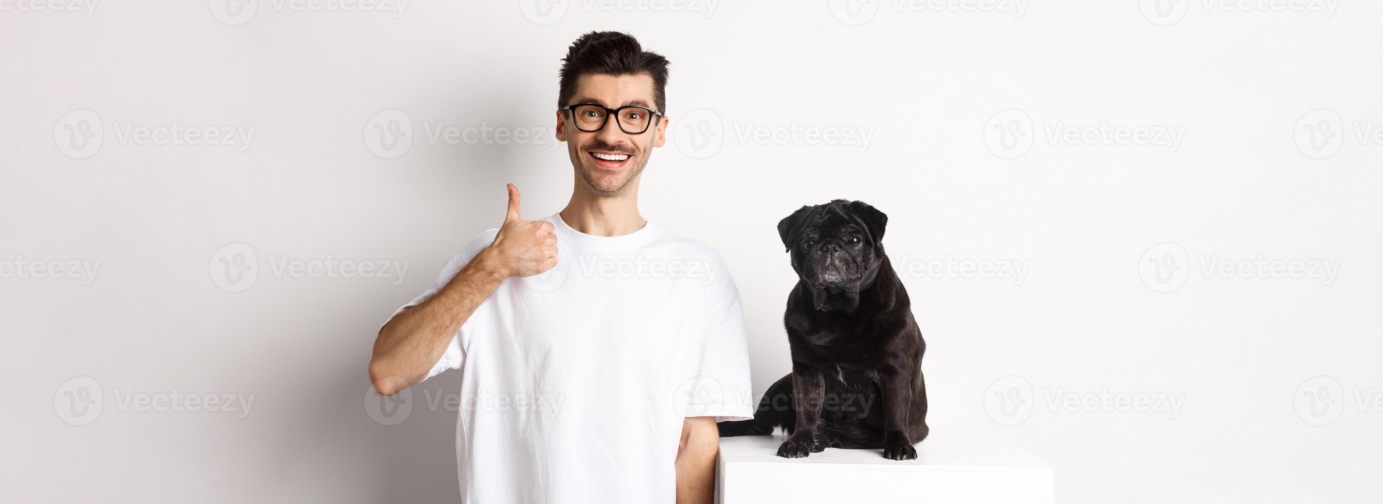 Image of dog owner and cute black pug looking at camera, man showing thumb-up in approval, recommending something, standing over white background photo