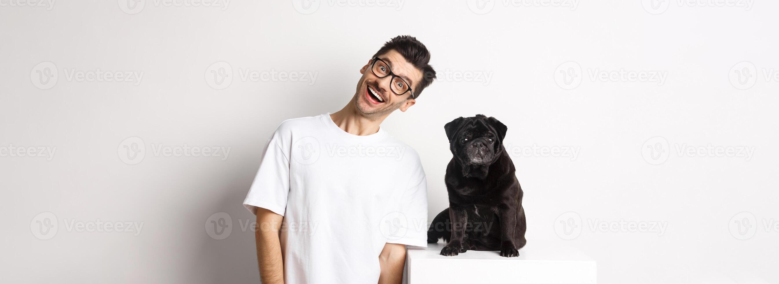 Image of handsome young man standing near cute black pug and smiling. Dog owner spending time with his pet, staring at camera happy, white background photo