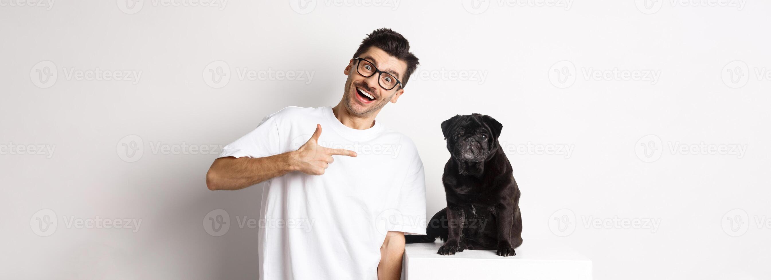 Happy young man showing his cute dog, pointing finger at black pug and smiling, standing over white background photo
