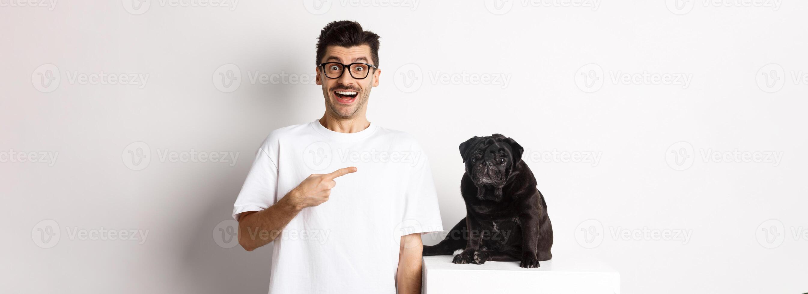 Happy young man showing his cute dog, pointing finger at black pug and smiling, standing over white background photo