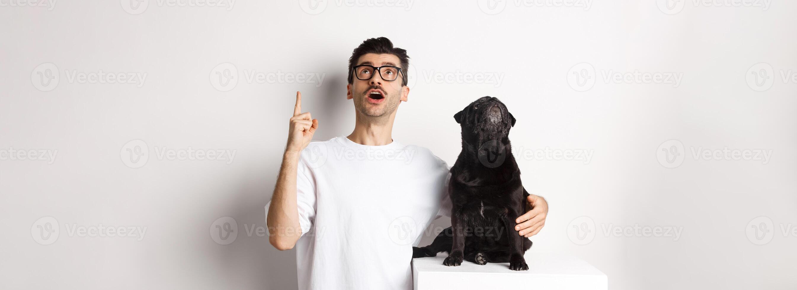 Image of happy young man and dog both looking up at promo, owner pointing finger at top, pug staring at loog, standing over white background photo