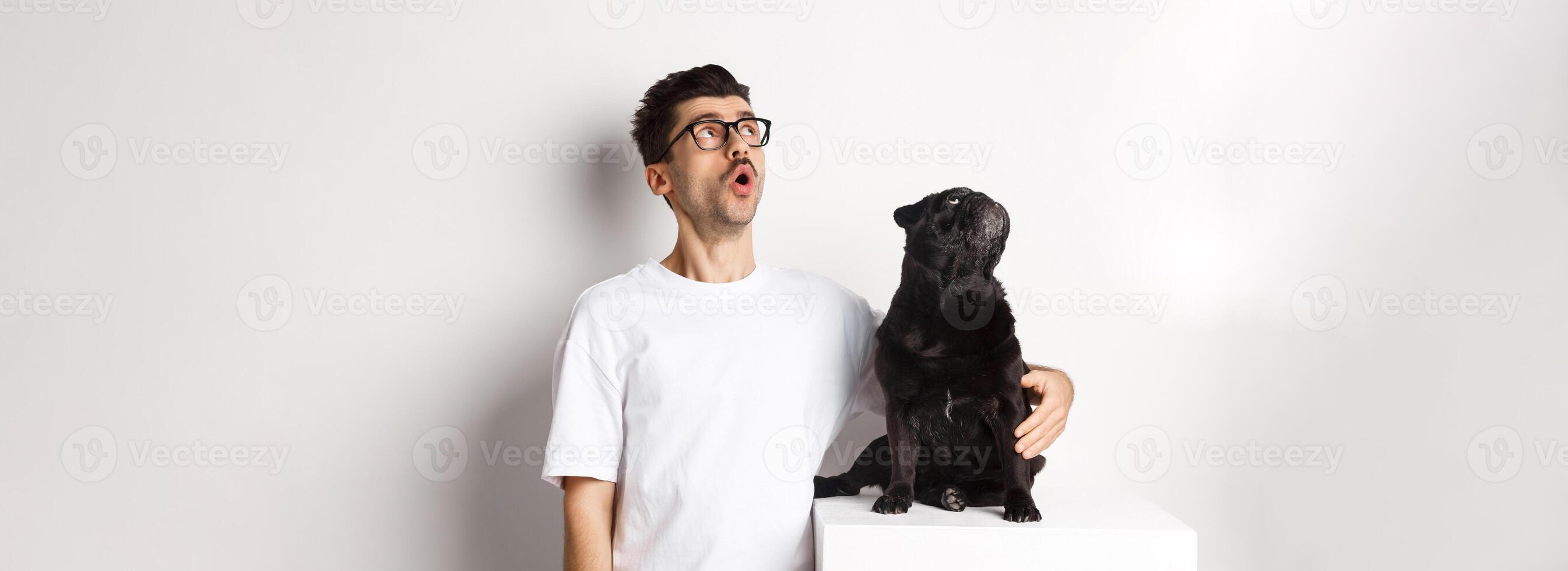 Amazed young man in glasses hugging his dog, pet owner and pug staring at upper left corner promo offer, standing over white background photo