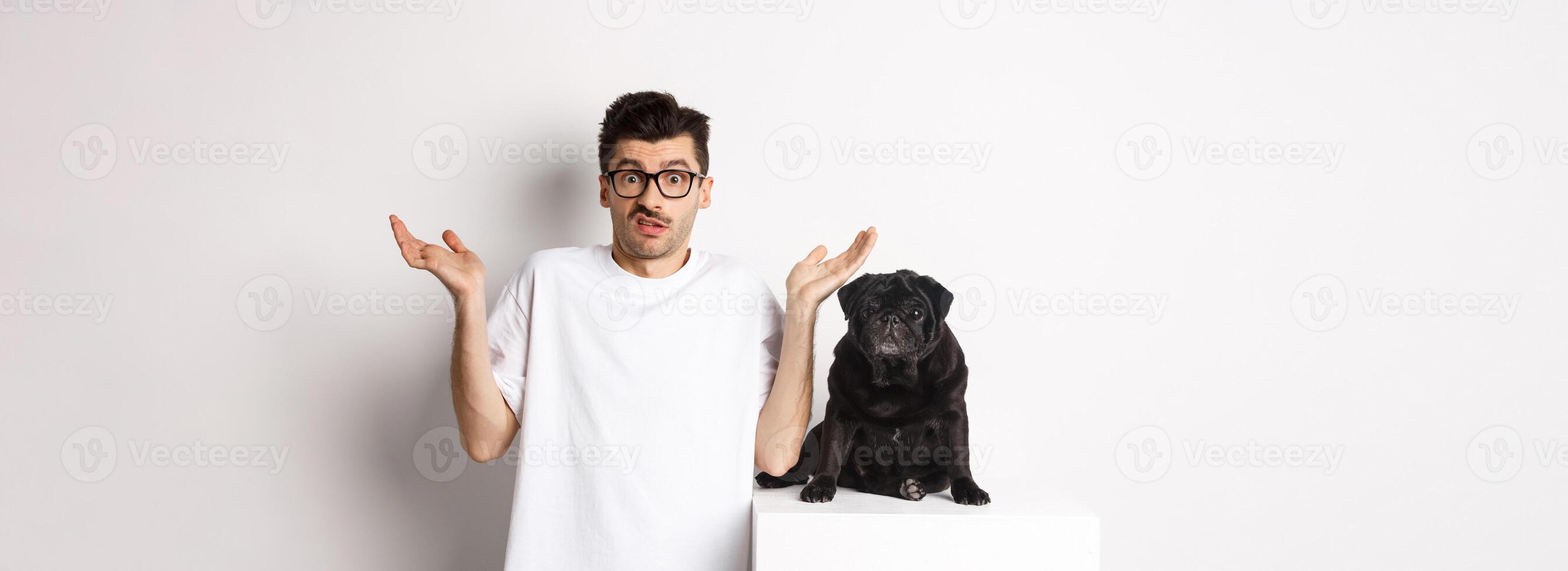 Image of confused man in glasses raising hands and shrugging complicated, standing near black pug dog over white background photo